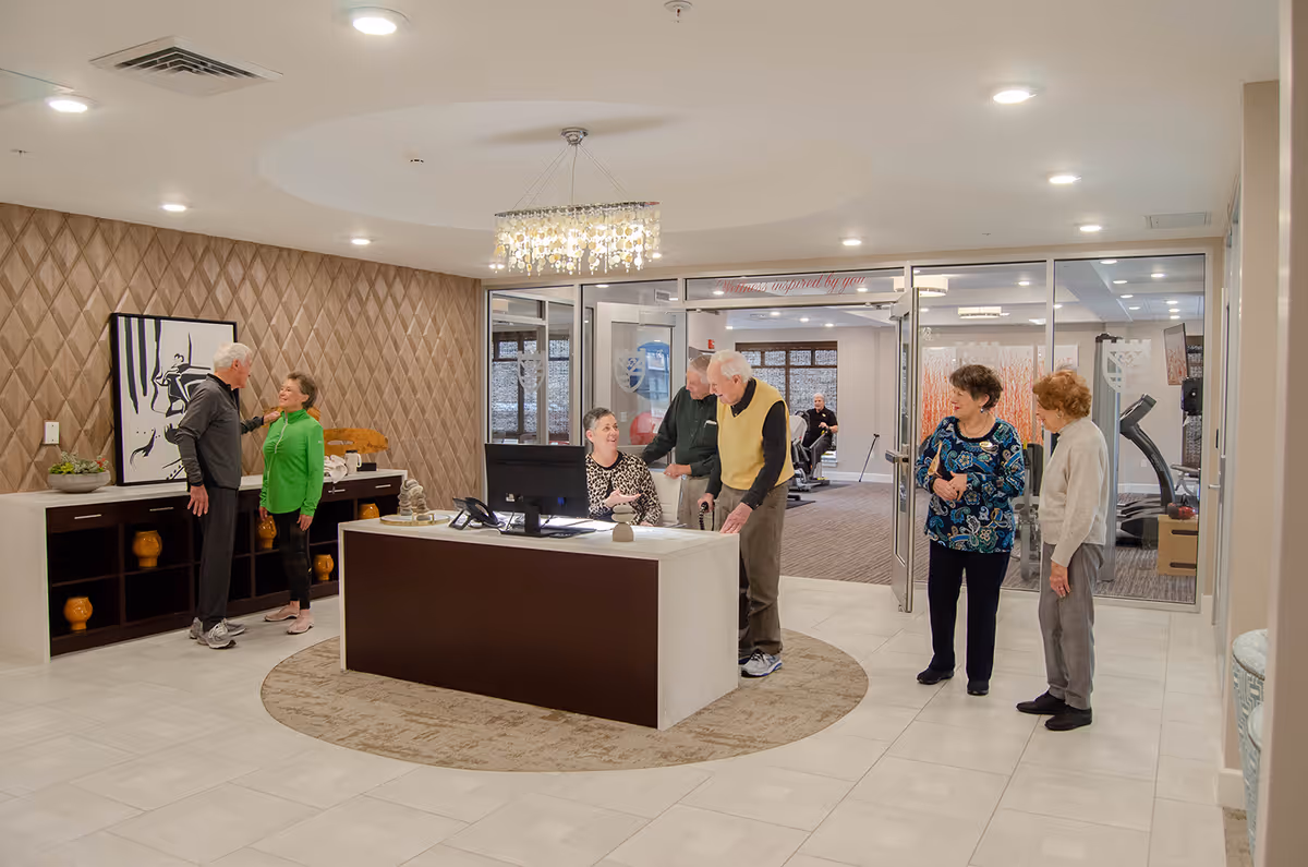 A senior living facility reception area with a front desk attended by a woman. Several elderly people are standing and interacting around the desk. The room has a modern design with a chandelier, patterned wallpaper, and a large framed artwork on the wall. Behind the reception area, there is a glass wall and door leading to a fitness room with exercise equipment.
