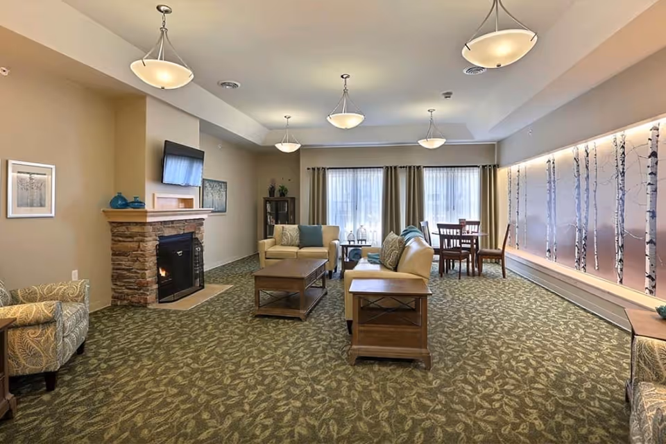 A spacious senior living common area with patterned green carpet, beige walls, and multiple ceiling lights. The room features a stone fireplace with a mounted TV above it, two beige sofas with blue and patterned cushions, a wooden coffee table, and side tables. In the background, there is a dining table with four chairs near large windows with curtains. The right wall has a decorative panel with birch tree designs illuminated by soft lighting.