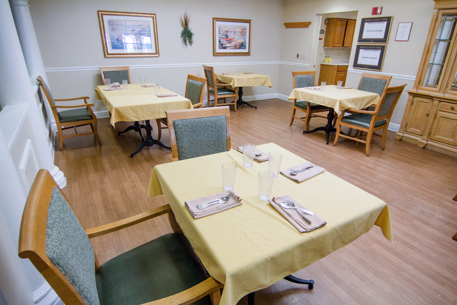 A dining room with several tables covered in yellow tablecloths, each set with napkins, utensils, and clear drinking glasses. The room has wooden chairs with green cushioned seats and backs, light-colored walls with framed artwork, and a wooden cabinet on the right side.