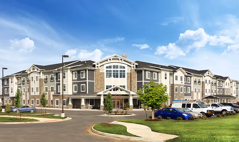 Front exterior of a three-story senior living community building with a porte-cochere entrance, parking lot and landscaped lawn under a blue sky.