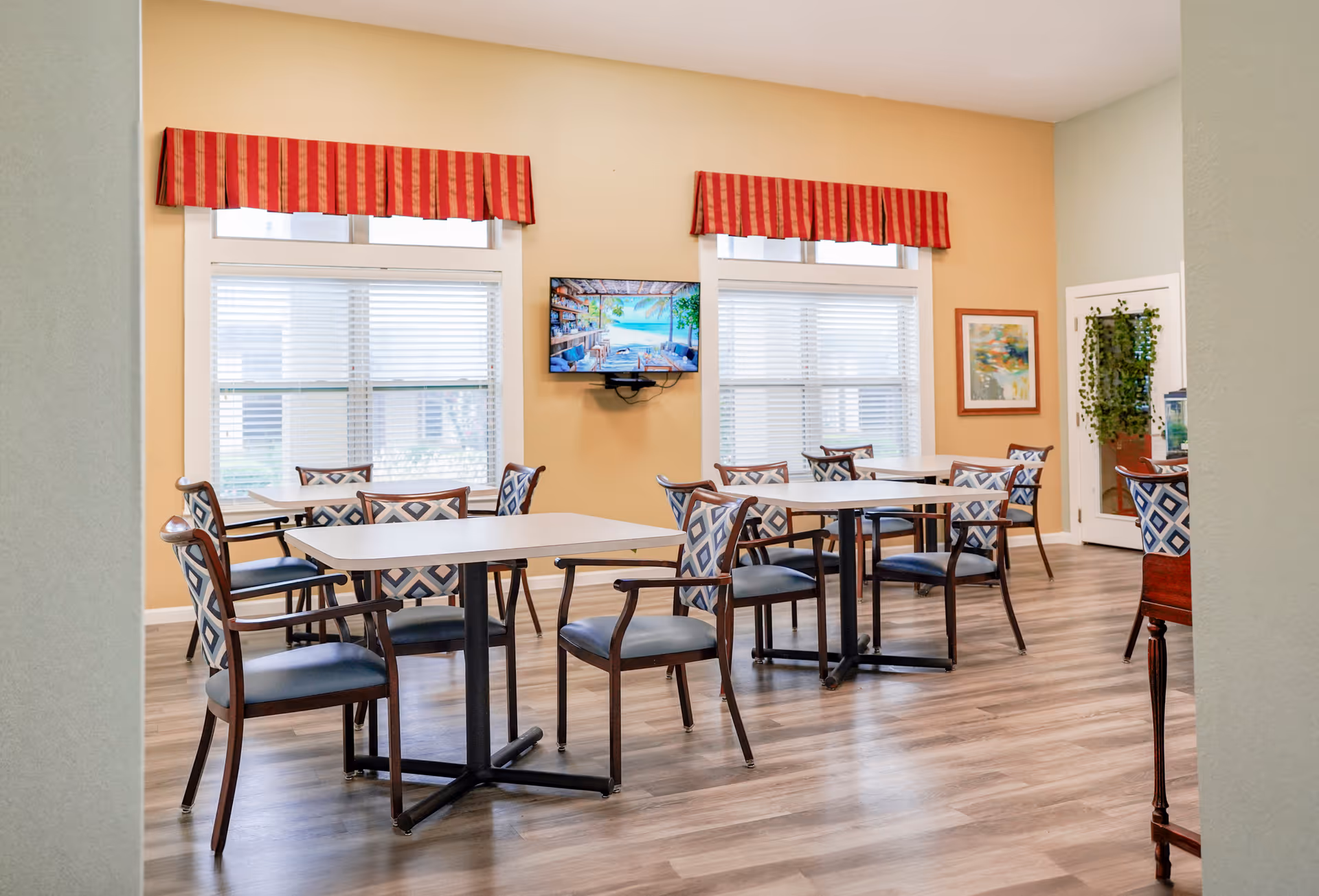A bright dining room with several tables and chairs arranged neatly. The chairs have blue and white patterned backs and dark wooden frames. Two large windows with white blinds and red striped valances let in natural light. A flat-screen TV is mounted on the wall between the windows, displaying a scenic beach image. There is a framed artwork on the wall and a door with a hanging green plant beside it. The floor is covered with light wood laminate.