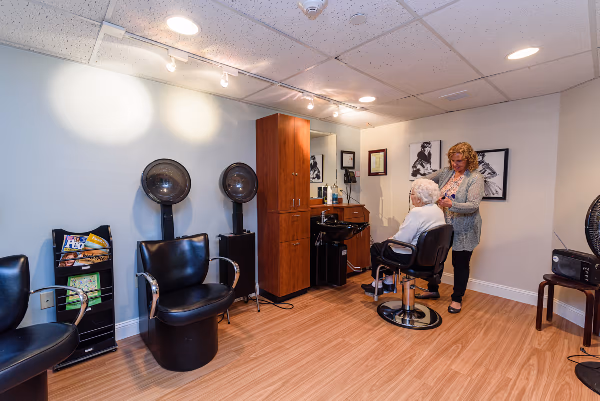 A hair salon area inside a senior living facility with wooden flooring and light-colored walls. There are two black salon chairs with hair dryers behind them, a wooden cabinet with a sink, and a stylist attending to an elderly woman seated in a salon chair. The walls have framed black and white pictures, and there is a magazine rack with magazines on the left side.