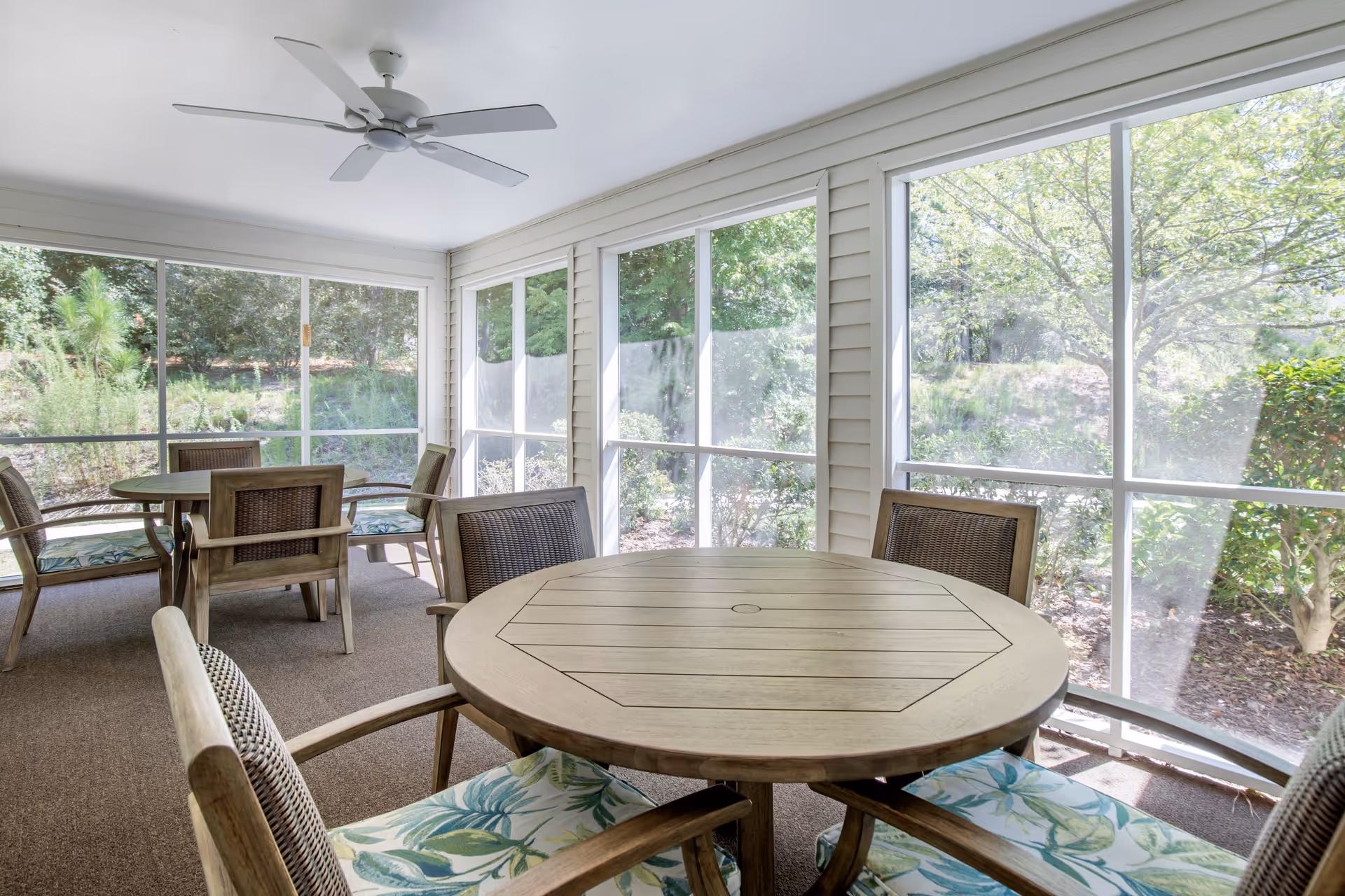 A bright enclosed patio area with large windows showing green trees and bushes outside. The space contains round wooden tables with cushioned chairs featuring a leafy pattern. A ceiling fan is mounted on the white ceiling.