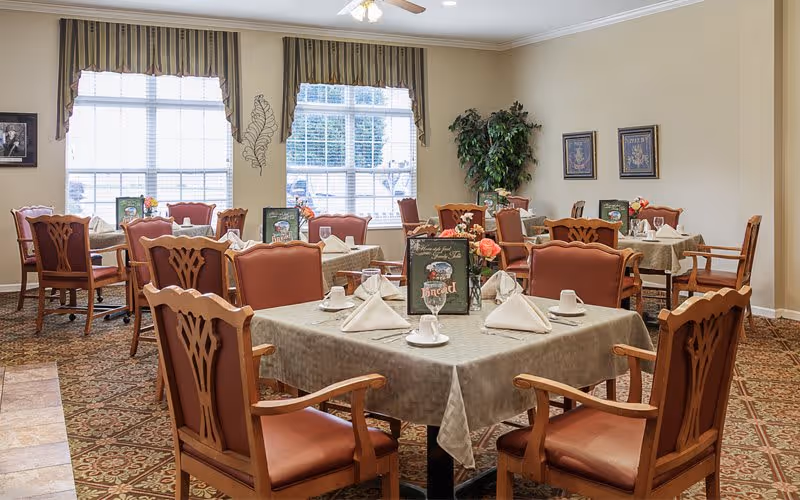 Interior view of the dining room at Bickford of Omaha Hickory, featuring tables set with napkins and menus, surrounded by comfortable chairs and large windows.