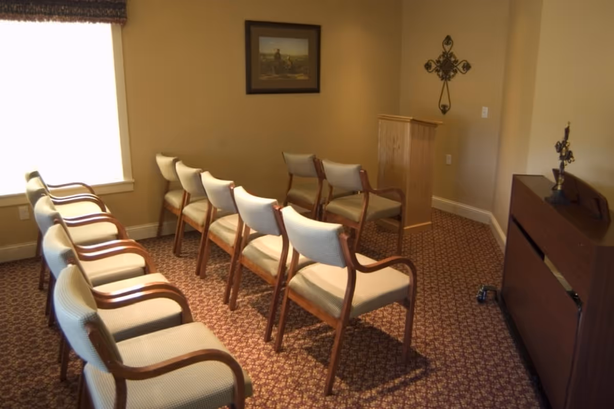 A small room with two rows of beige cushioned chairs with wooden armrests facing a wooden podium. The room has beige walls, a patterned carpet, a window with a dark valance, a framed picture on the wall, and decorative crosses on the walls and a cabinet.