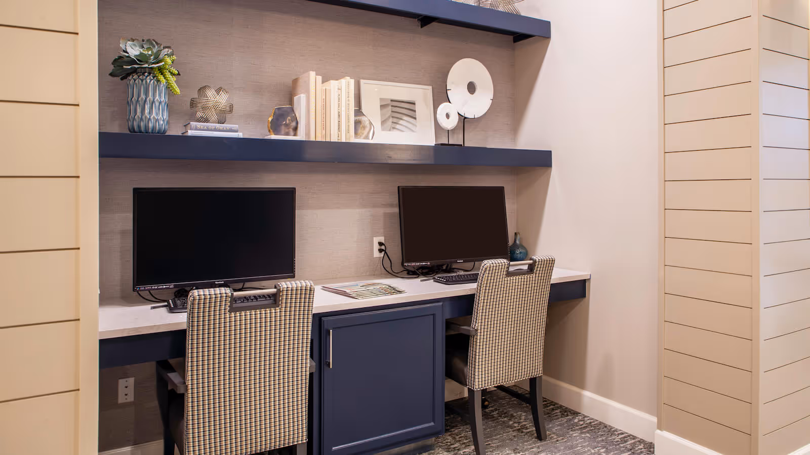 Two computer workstations with monitors and upholstered chairs built into a wall desk with shelves and decorative items.