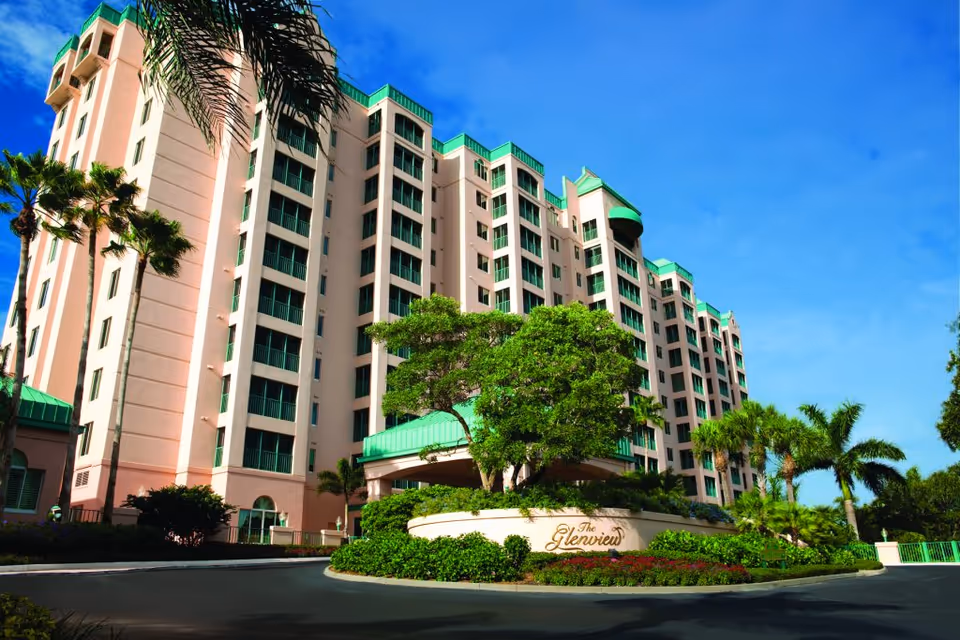 Exterior view of a large multi-story senior living facility named The Glenview, surrounded by palm trees and greenery under a clear blue sky.