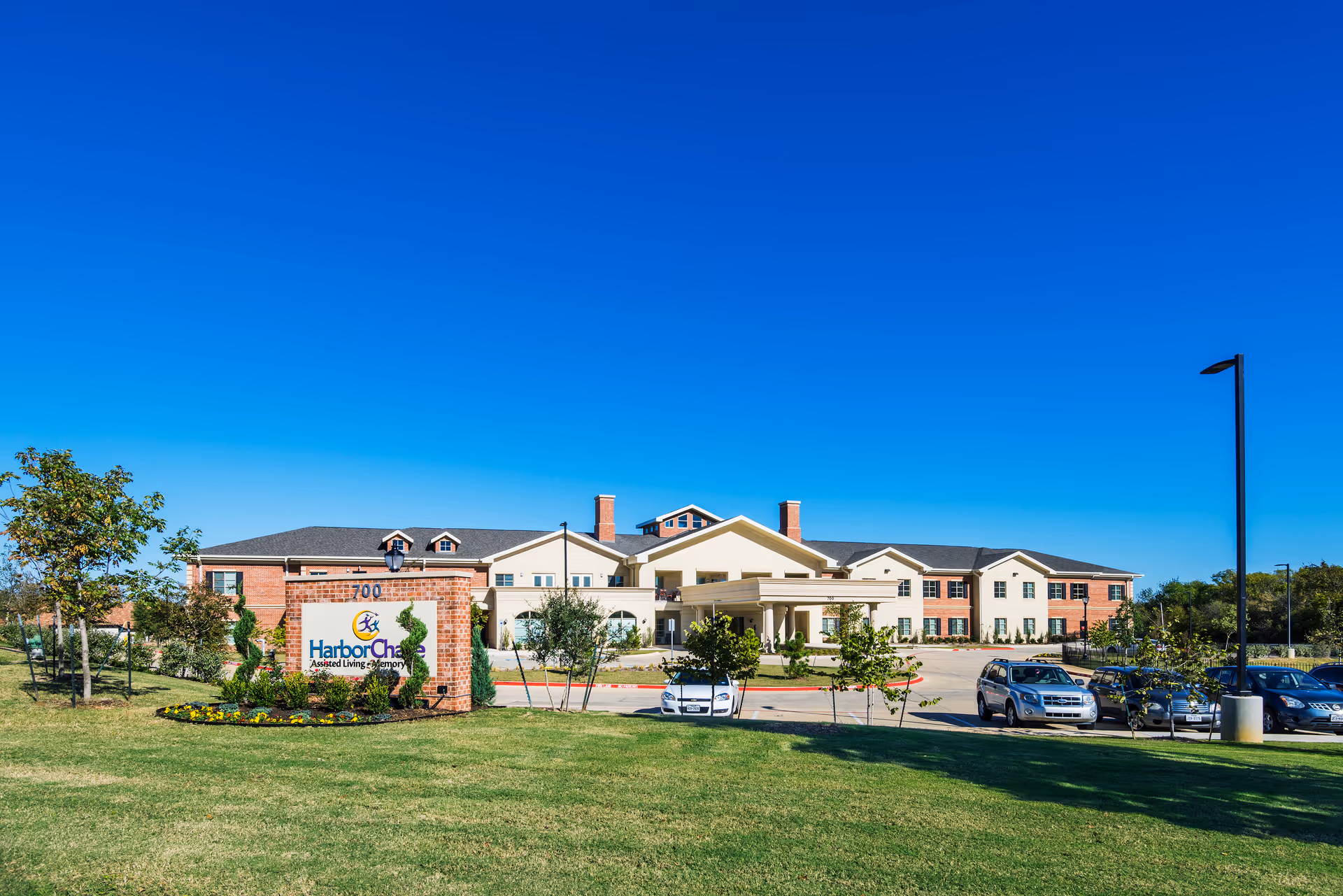 Exterior view of HarborChase of Southlake assisted living and memory care facility with a clear blue sky, green lawn, trees, and parked cars in front of the building.