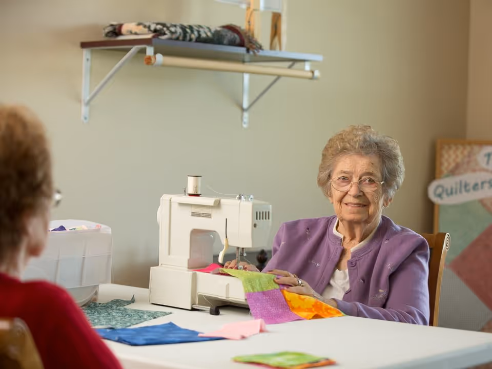 An elderly woman wearing glasses and a purple cardigan is sitting at a table using a sewing machine to work on a colorful quilt. Another person is partially visible in the foreground. The room has a shelf with folded blankets and a sign in the background that reads 'Quilters'.