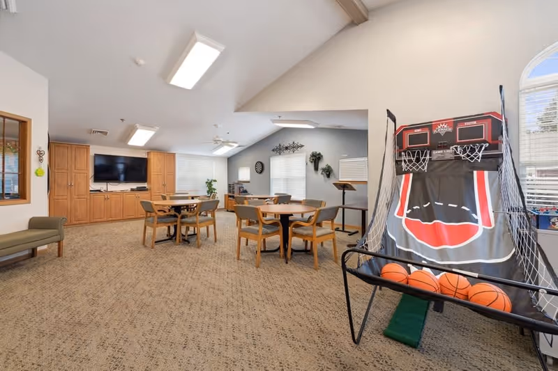 A spacious common area with carpeted floor, featuring round tables with chairs, a large flat-screen TV mounted on a wooden cabinet, and a basketball arcade game with several basketballs. The room has large windows with blinds and a high ceiling with skylights.