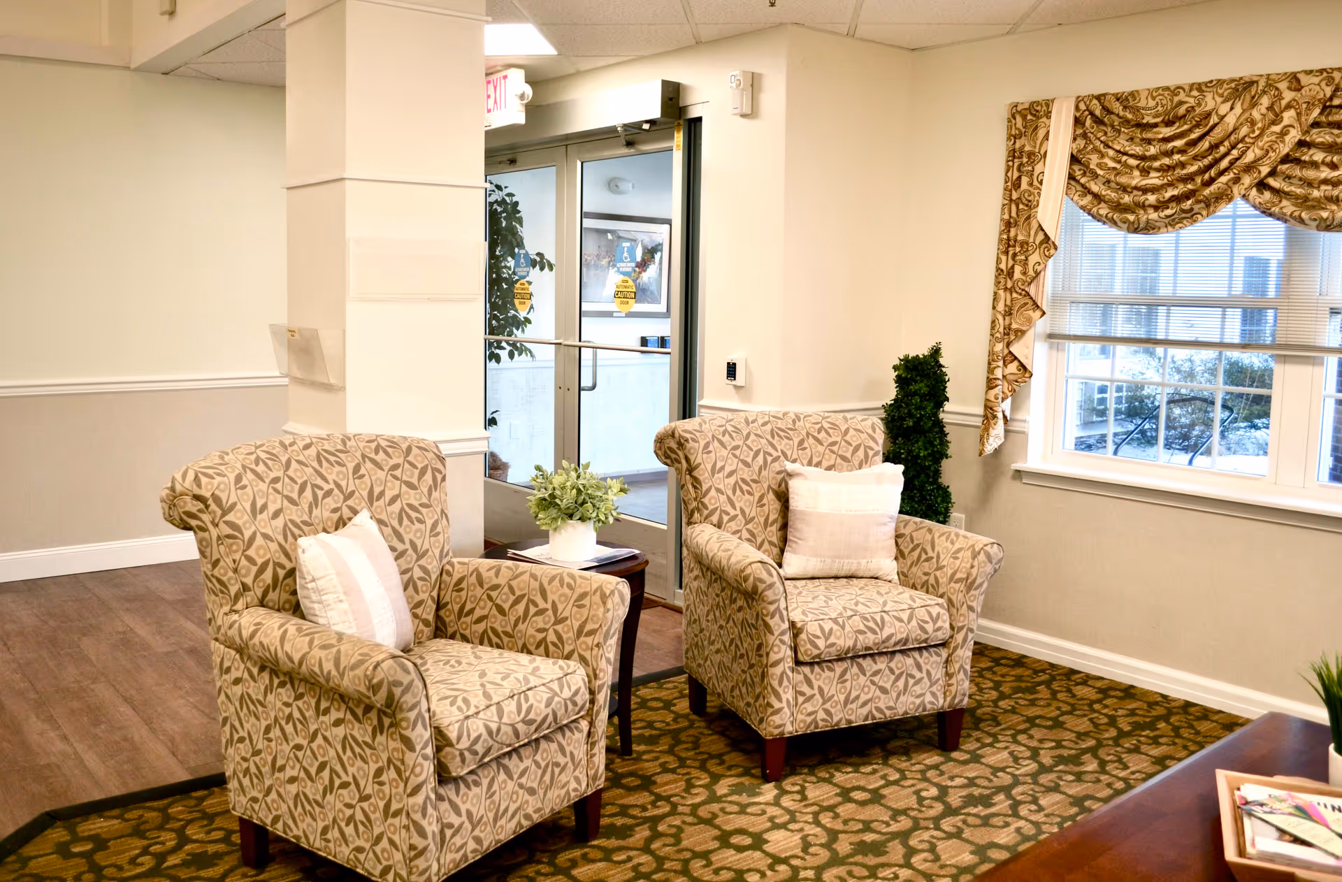 A cozy seating area in a senior living facility with two patterned armchairs each with a white pillow, a small wooden side table with a potted plant, a window with decorative curtains, and a glass door leading outside.