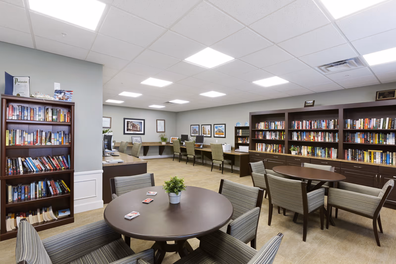 Bright community library/activity room with round tables, upholstered chairs, bookshelves and computer workstations.