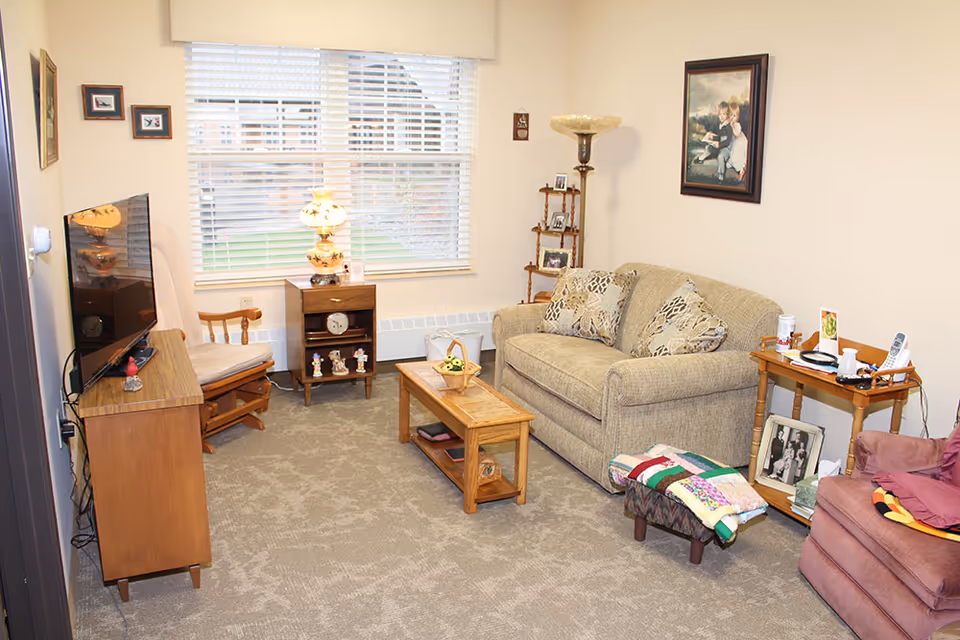 Cozy senior living community sitting room with a sofa, armchairs, TV, coffee table and window with blinds.