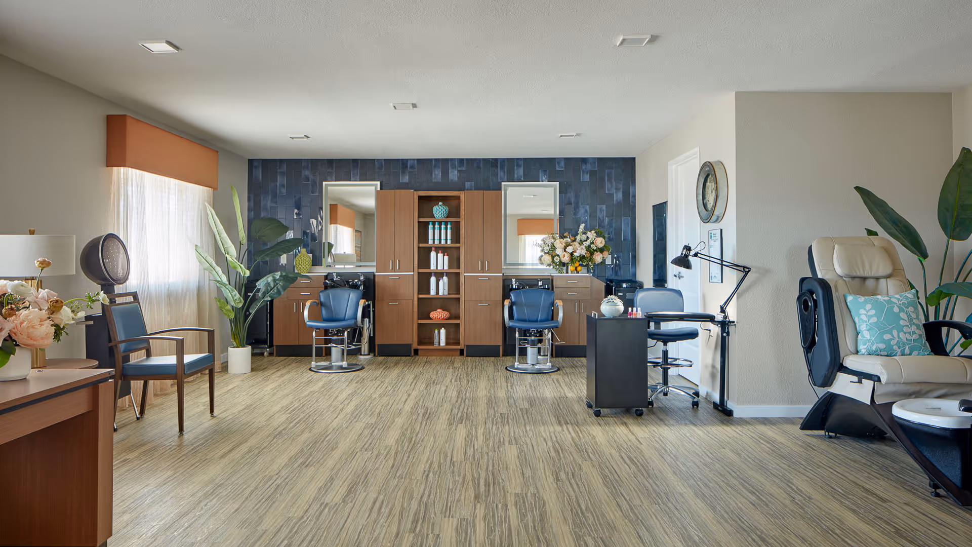 A bright and spacious salon area with two blue salon chairs in front of mirrors and wooden cabinets stocked with hair care products. There are plants and floral arrangements on either side, a manicure station with a black rolling chair and lamp, and a comfortable cream-colored massage chair with a teal pillow on the right. The room has light wood flooring and neutral-colored walls.