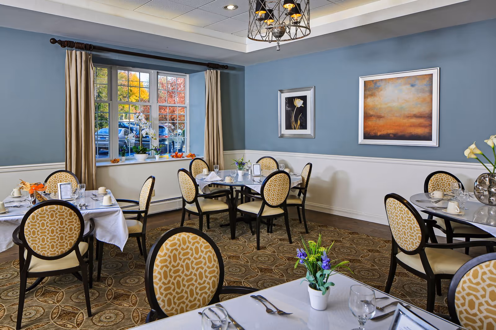 A dining room with several round tables covered with white tablecloths, each set with cups, glasses, and silverware. The chairs have patterned yellow upholstery. The walls are painted blue with white wainscoting, and there are two framed paintings on the wall. A window with beige curtains shows a view of parked cars and autumn trees outside. A decorative chandelier hangs from the ceiling.