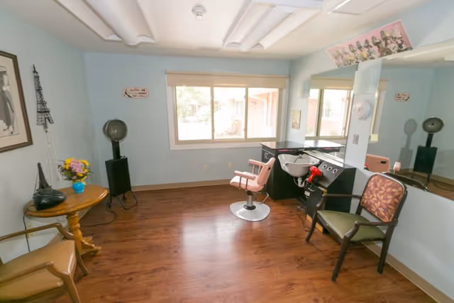 A small hair salon room with light blue walls and wooden flooring. The room contains a pink salon chair in front of a black hair washing station with a white sink. There is a large mirror on the right wall, a vintage hair dryer on a stand in the corner, and two chairs with patterned cushions. A small round wooden table holds a vase with flowers and a decorative Eiffel Tower. A window lets in natural light.