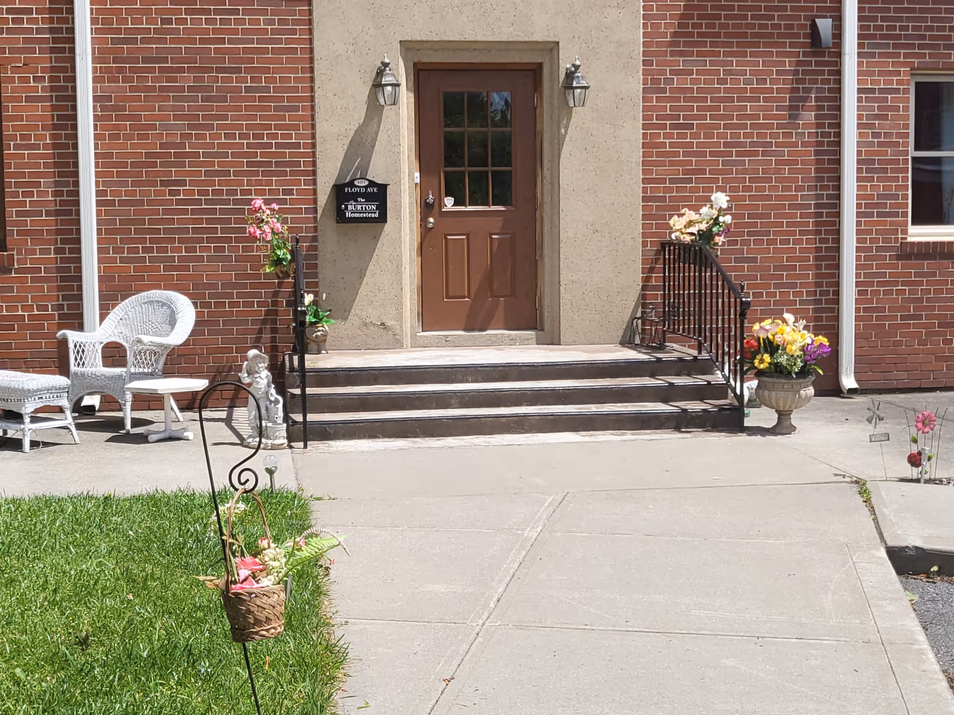Entrance to The New Burton Homestead featuring a brown door with glass panels, brick walls, concrete steps with black railings on both sides, white wicker chair and table on the left, and flower pots with colorful flowers on both sides of the steps and near the walkway.