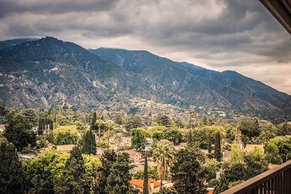 View of a residential neighborhood with many trees and houses in the foreground, and large mountains under a cloudy sky in the background, seen from a balcony with a railing on the right side.