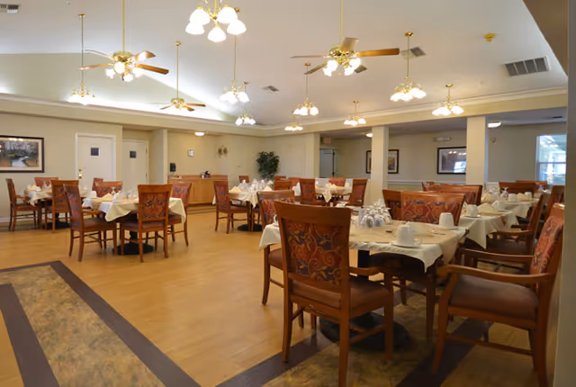 A spacious dining room with multiple wooden tables and chairs arranged neatly. Each table is covered with a white tablecloth and set with cups, saucers, and napkins. The room has wooden flooring, ceiling fans with lights, and soft overhead lighting fixtures. There are framed pictures on the walls and some plants in the background.