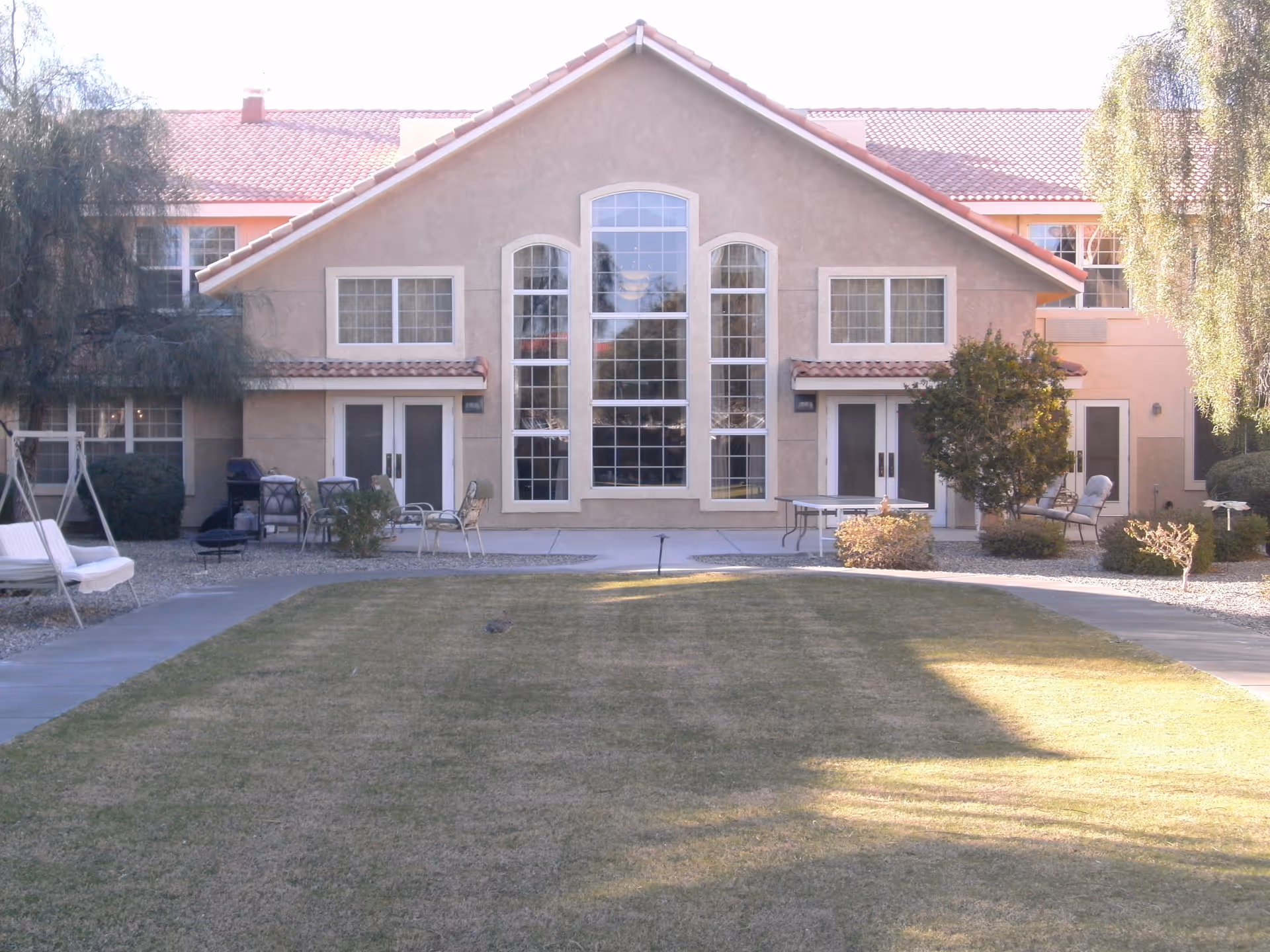 Outdoor view of a senior living facility building with a large central window and two smaller windows on either side. The building has a beige stucco exterior with a red tile roof. In front of the building is a grassy lawn with a paved walkway, patio furniture, a swing, and some bushes and trees.