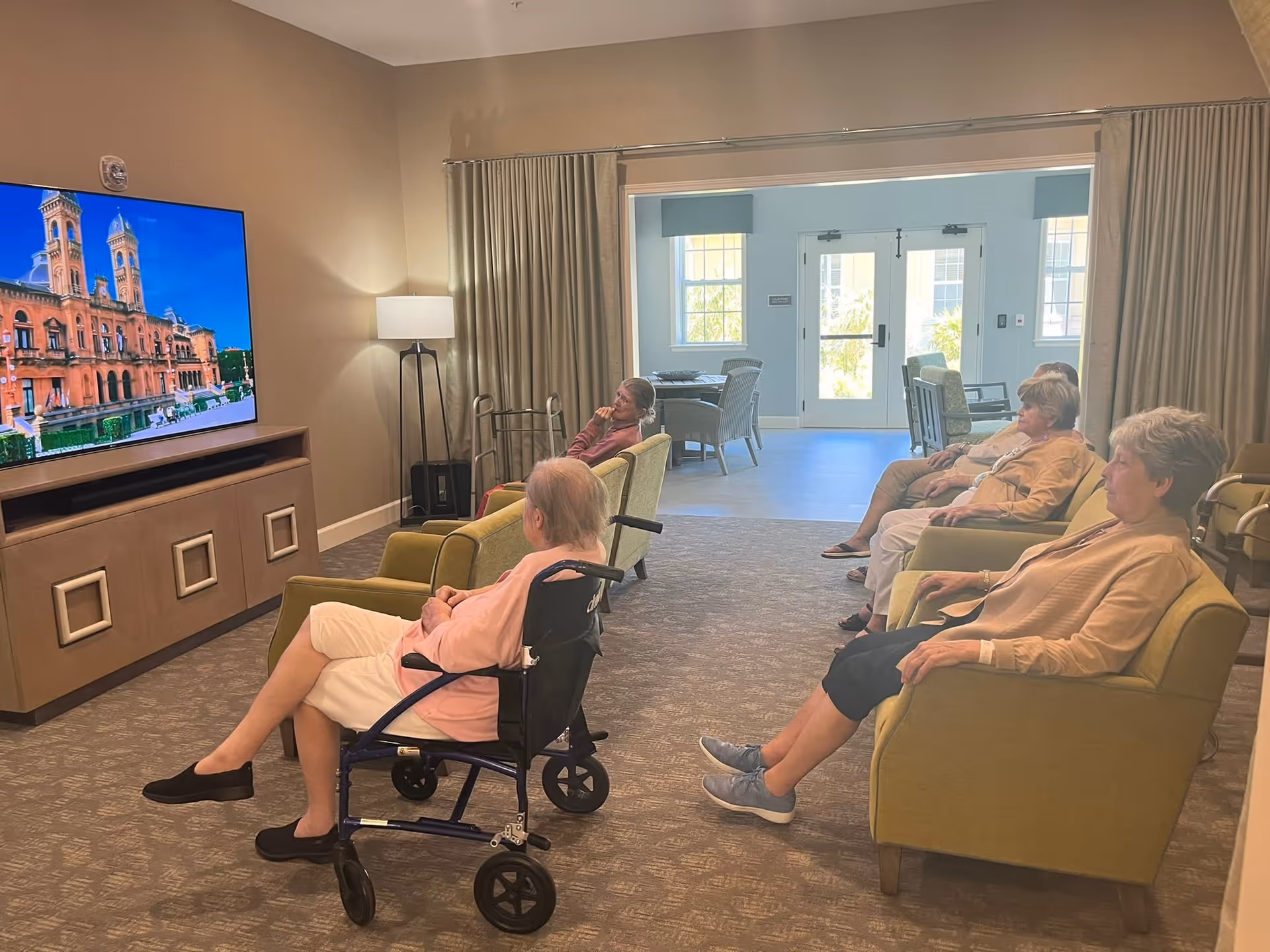 A group of elderly women sitting in a living room area watching a large flat-screen TV mounted on the wall. The room has beige walls, carpeted floor, and comfortable armchairs. There is a floor lamp and a walker near the TV stand. The background shows a bright area with windows and a door leading outside.