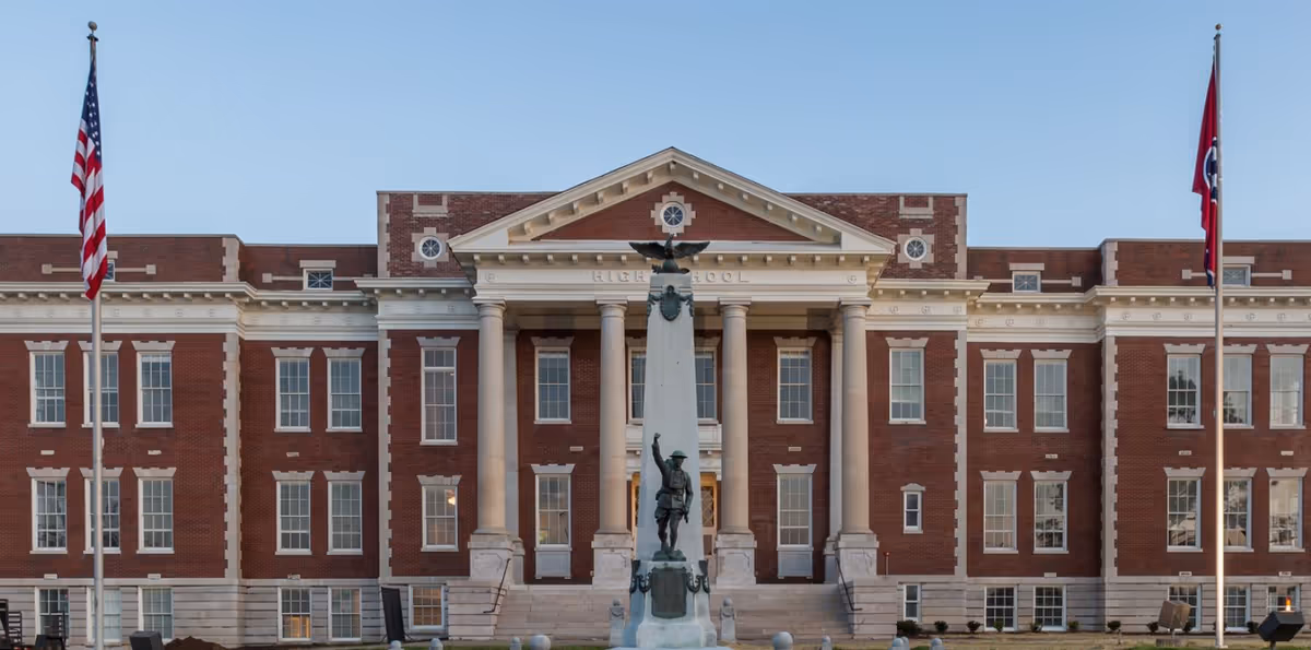 Front exterior view of a large brick building with classical architectural features including columns and a triangular pediment. In front of the building is a tall monument with a statue of a soldier and an eagle on top. Two flagpoles with flags flank the monument on either side.