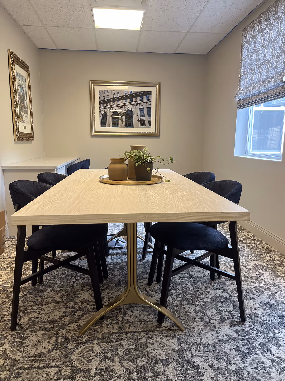 A small dining area with a light wooden table and six black cushioned chairs arranged around it. On the table, there is a decorative tray holding two brown vases and a small potted plant. The room has beige walls with two framed pictures hanging, a window with a patterned Roman shade, and a patterned carpeted floor.