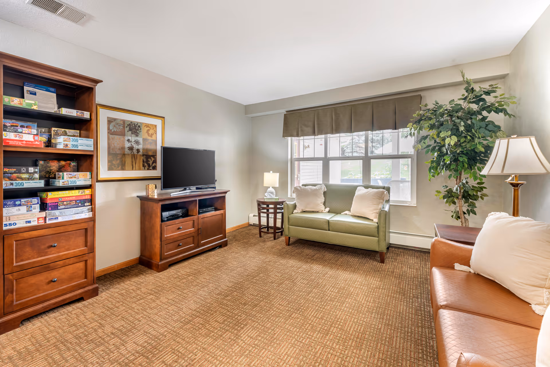 A cozy living room with a green loveseat and a brown leather sofa, both with white pillows. There is a wooden TV stand with a flat-screen TV and a tall wooden bookshelf filled with board games. A large window with a valance lets in natural light, and there are two table lamps and a tall potted plant adding to the decor.