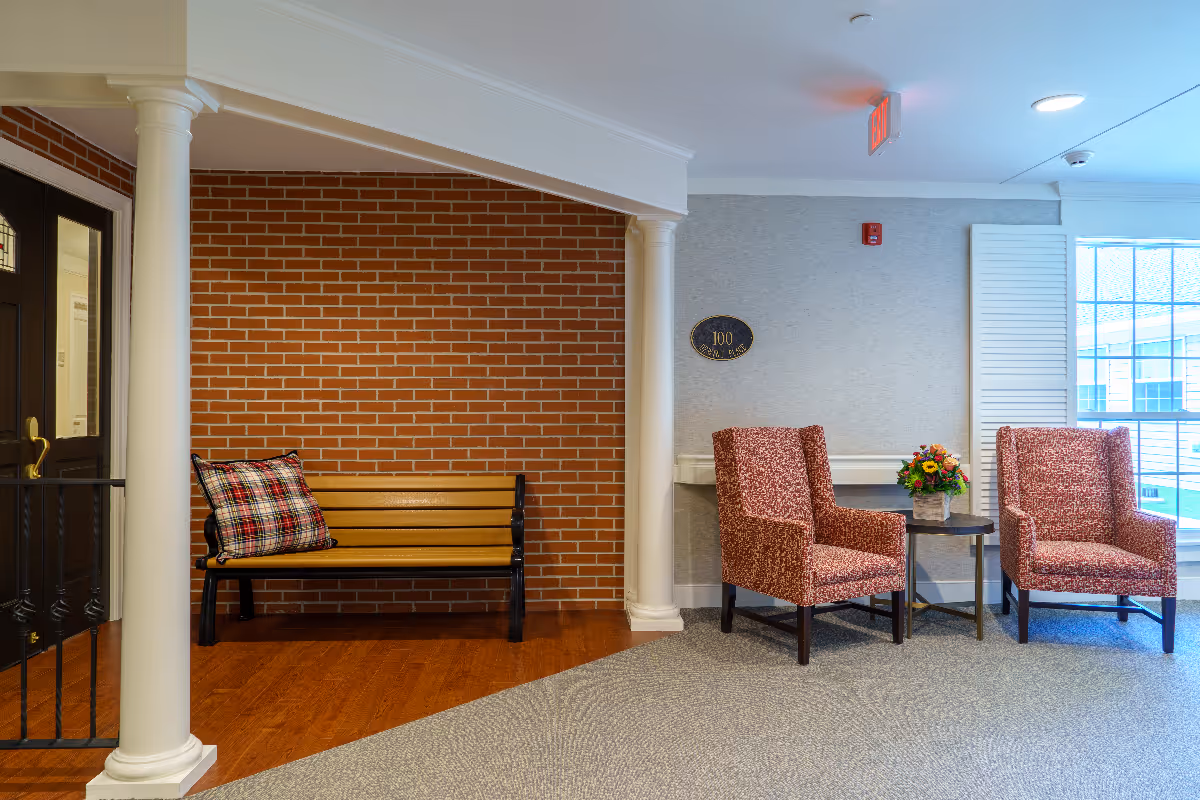 Interior seating area with two patterned armchairs and a small round table with a flower arrangement between them. To the left, there is a wooden bench with a plaid cushion against a red brick wall. The floor is a combination of carpet and wood, and there are white columns and a door visible on the left side.