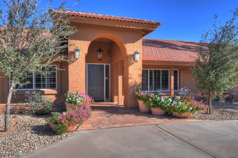 Front exterior view of a single-story building with a terracotta tiled roof and an arched entryway. The entrance has a gray door flanked by two wall-mounted lantern lights. There are several potted plants with colorful flowers on either side of the walkway leading to the door. Small trees and desert landscaping with rocks surround the building under a clear blue sky.