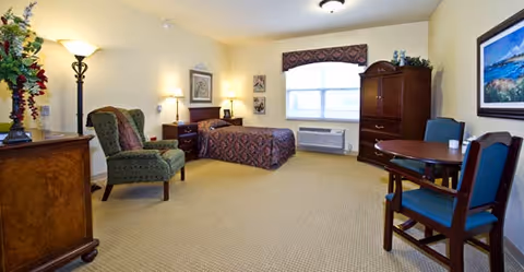 A spacious bedroom in Cedar Creek Health Campus featuring a single bed with patterned bedding, a green upholstered armchair, a wooden dresser with a lamp and floral arrangement, a wooden armoire, a round table with two chairs, and a window with a valance letting in natural light.