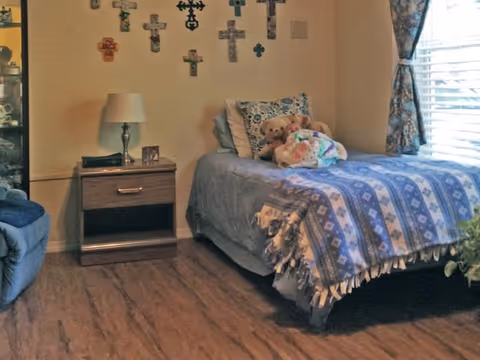 A cozy bedroom with a single bed covered in a blue patterned blanket and pillows, including two teddy bears and a colorful stuffed animal on the bed. A wooden nightstand with a lamp and a small framed photo is next to the bed. The wall above the bed is decorated with various crosses. A window with floral curtains allows natural light into the room, and part of a blue armchair is visible on the left side.