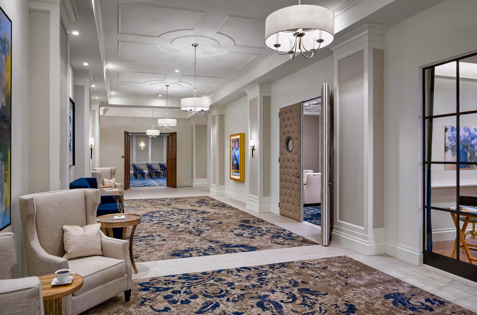 A spacious, elegantly decorated hallway with beige and blue patterned carpet, beige armchairs with small wooden side tables, modern chandeliers hanging from a white ceiling with decorative molding, and a partially open padded door leading to a room with blue carpet and chairs.