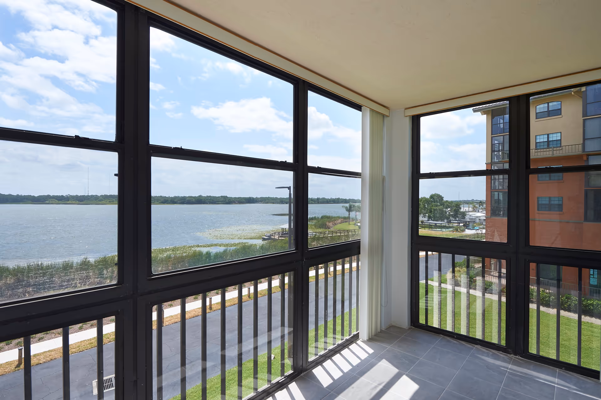 A bright enclosed balcony with large black-framed windows overlooking a lake and greenery. The balcony has tiled flooring and vertical blinds partially pulled to the side. Outside, there is a paved path, grassy areas, and a multi-story building visible to the right.