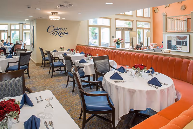 Interior view of a dining room named Cypress Room with round and rectangular tables covered with white tablecloths, set with blue napkins, silverware, and glasses. The room features orange cushioned bench seating along the wall, wooden chairs with blue cushions, and large windows letting in natural light. A few people are seated at tables in the background.