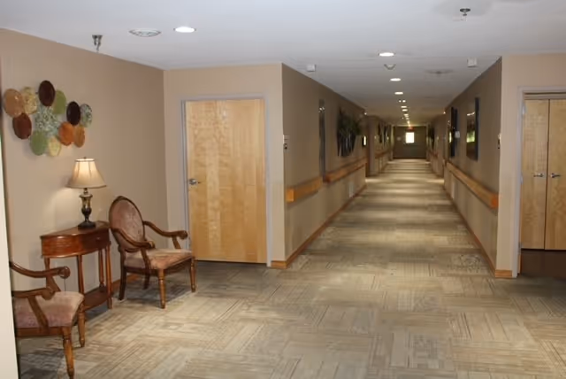 Long, well-lit interior hallway of a senior living facility with chairs, a side table and handrails along the walls.