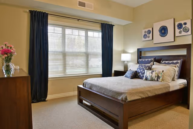A well-lit bedroom with a large window covered by dark curtains and blinds. The room features a wooden bed with a beige bedspread and multiple decorative pillows. There is a wooden dresser with a vase of pink flowers on the left side and a nightstand with a lamp on the right side. Three framed abstract artworks hang above the bed.