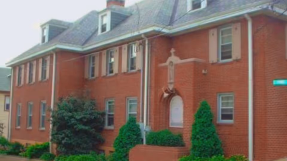 Two-story red brick building front with shrubs, dormer windows, and a decorative arched entry.
