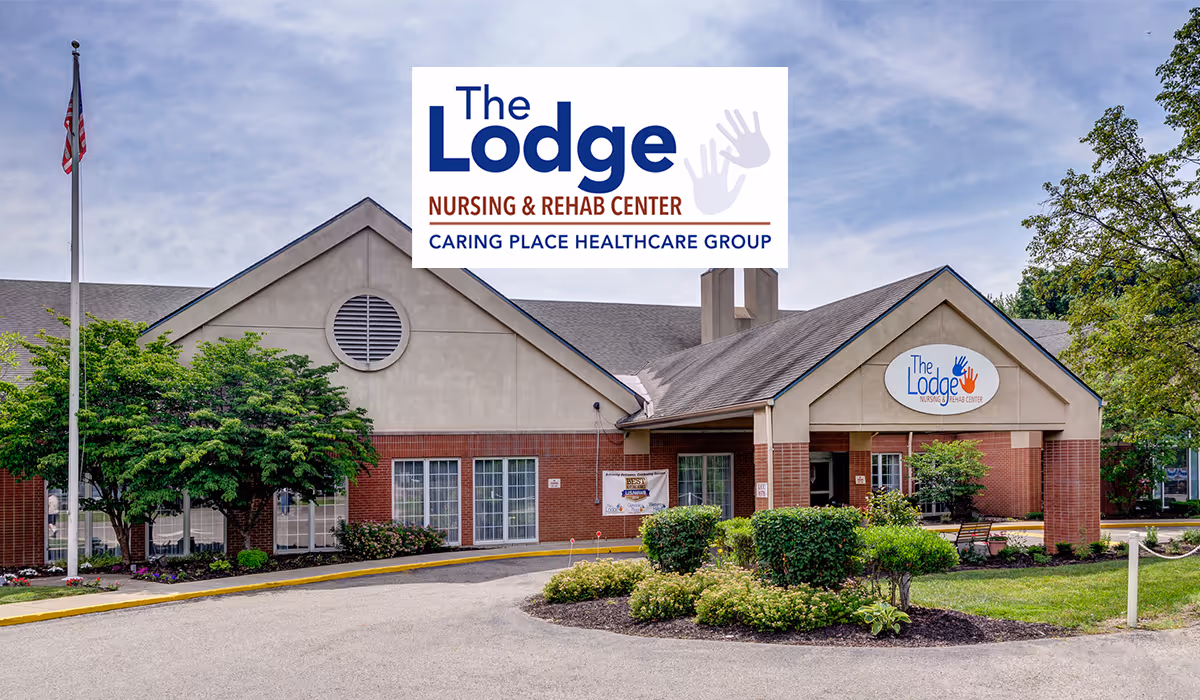 Exterior view of The Lodge Nursing & Rehab Center building with a driveway, landscaped bushes, trees, and an American flag on a flagpole under a partly cloudy sky.