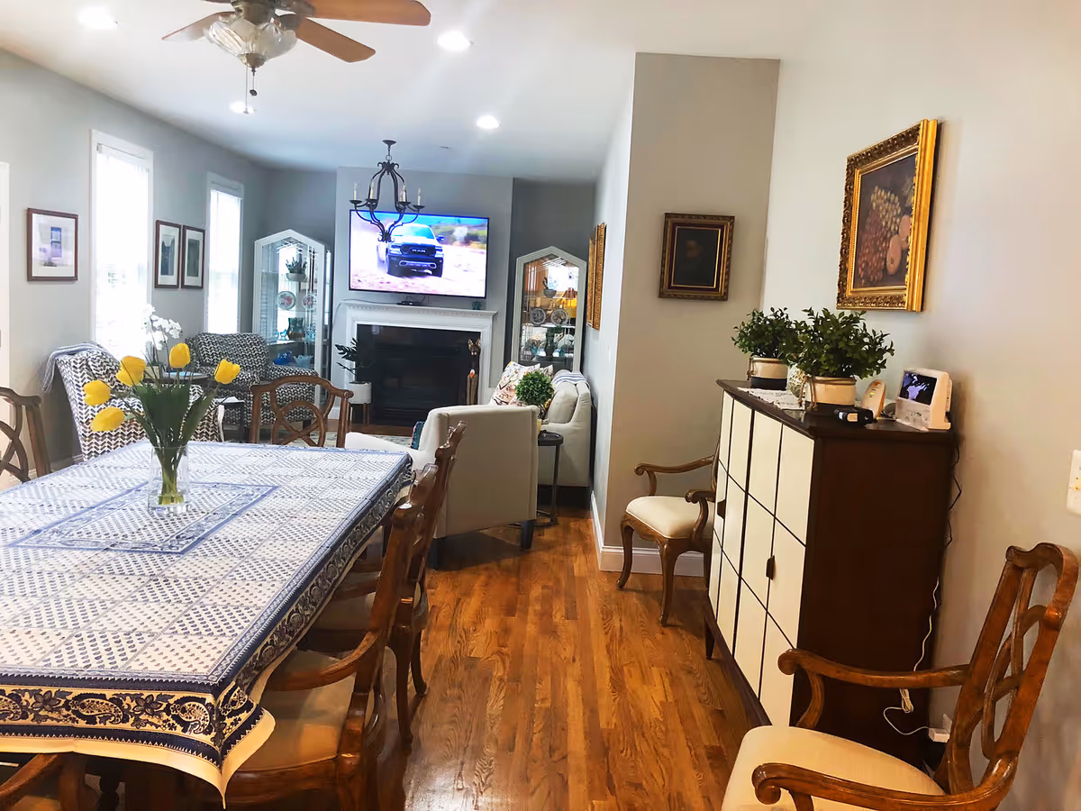Dining room with blue patterned tablecloth, tulip centerpiece, and adjacent living area with TV.