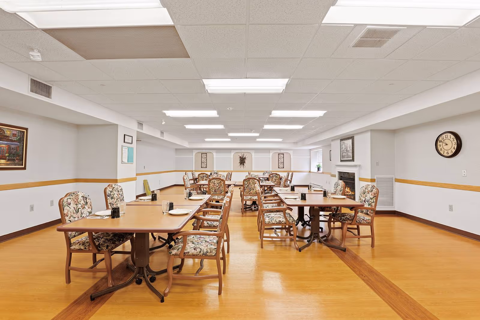 A senior living facility dining room with multiple wooden tables and floral upholstered chairs. The tables are set with plates, glasses, and utensils. The room has a light-colored ceiling with fluorescent lights, light walls with a wooden chair rail, a wall clock, framed artwork, and a fireplace on the right side.