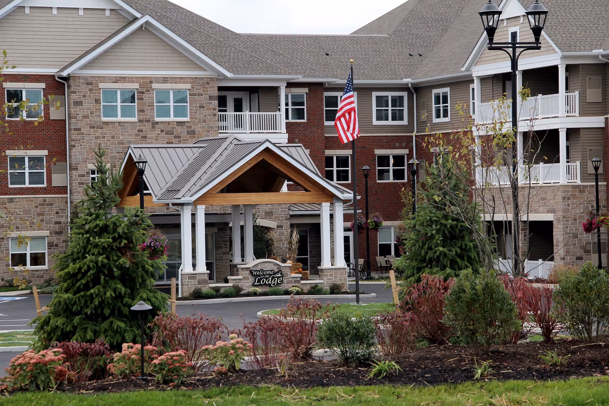 Exterior view of a senior living community building with a covered entrance labeled 'Welcome to the Lodge'. The building features a mix of stone and brick facade with multiple windows and balconies. There are landscaped bushes and trees in the foreground, an American flag on a pole near the entrance, and several lamp posts around the driveway.