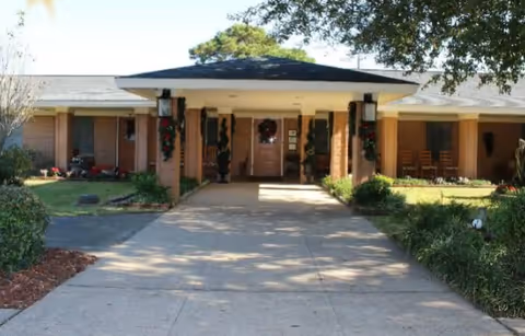 Front exterior view of a single-story assisted living facility with a covered entrance supported by columns decorated with holiday wreaths and greenery. There are bushes and trees around the building, and a concrete driveway leads up to the entrance.