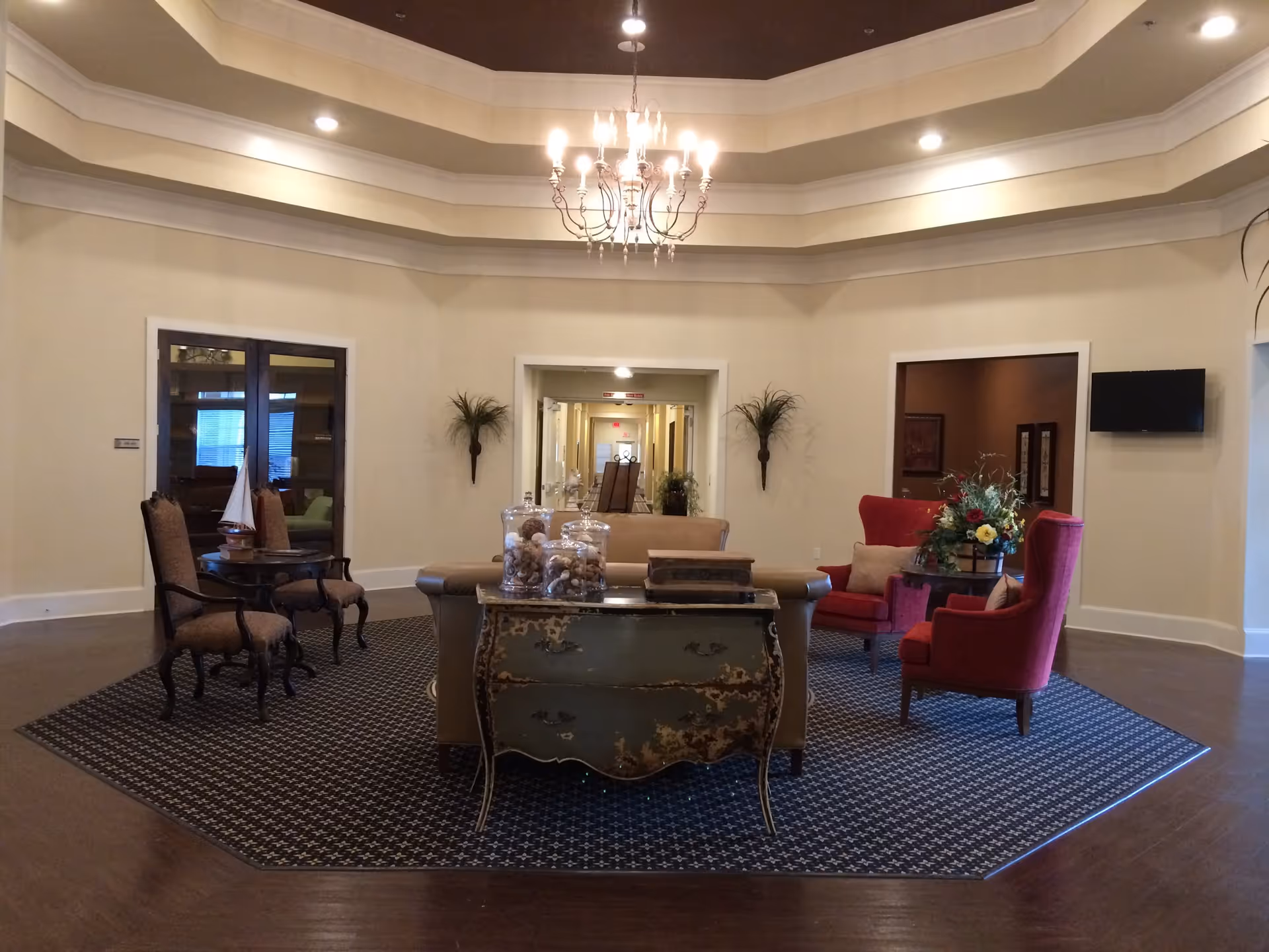 Well-lit senior living facility lobby with a chandelier, central seating area of armchairs and a sofa on a patterned rug and decorative console table.