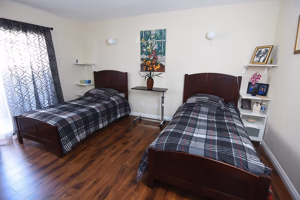 A senior care facility bedroom with two single beds featuring dark wooden frames and plaid bedding. Between the beds is a small black table with a vase of flowers. The room has wooden flooring, light-colored walls, and a window with patterned curtains on the left. Shelves on the walls hold framed photos, a small plant, and other decorative items.