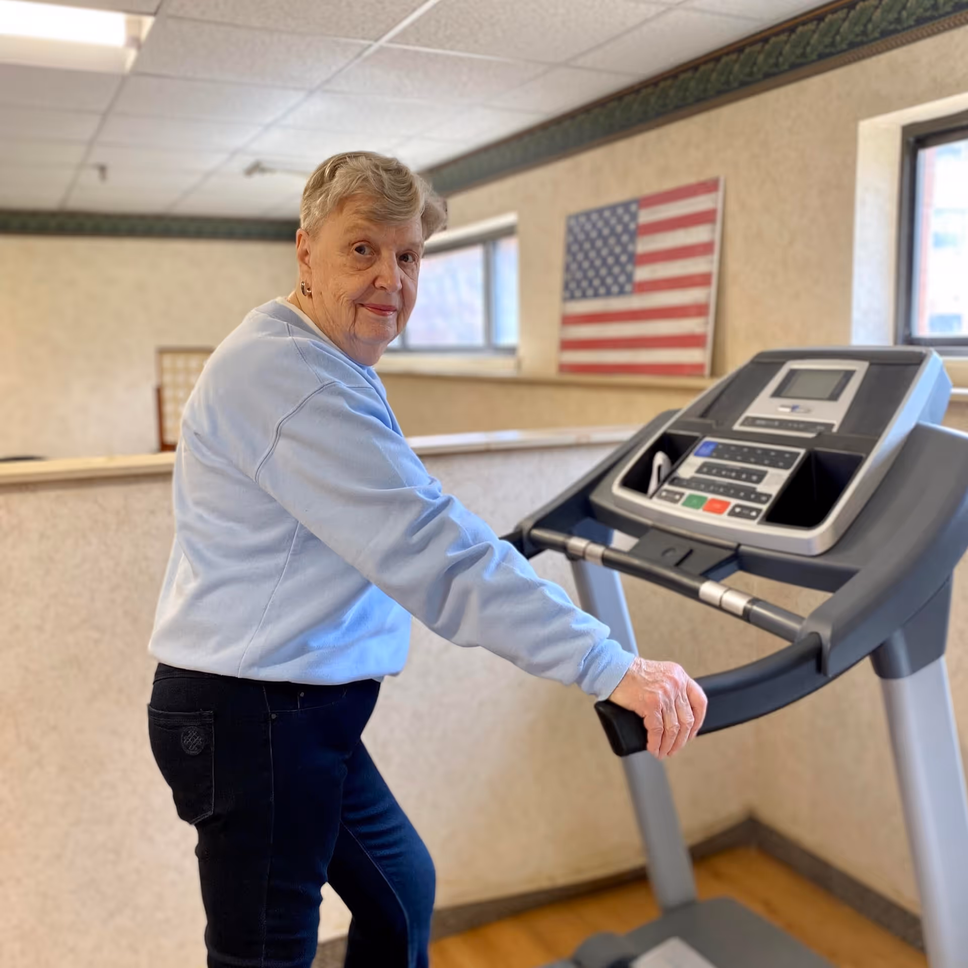 An elderly woman in a light blue sweatshirt and dark pants is using a treadmill in an indoor exercise area. Behind her, there is an American flag mounted on the wall and windows letting in natural light.