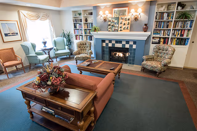 A cozy living room with a fireplace, surrounded by various upholstered chairs and a sofa. There are built-in bookshelves filled with books and plants on either side of the fireplace. A wooden coffee table and side table with a floral arrangement are also present. The room is well-lit with wall sconces and natural light from a window with draped curtains.