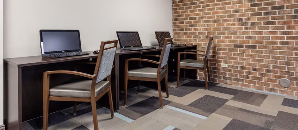 A small computer workstation area with three desktop computers on a dark wooden desk against a brick wall. Each computer has a wooden chair with a cushioned seat and backrest. The floor is carpeted with a patterned design in shades of gray and beige.