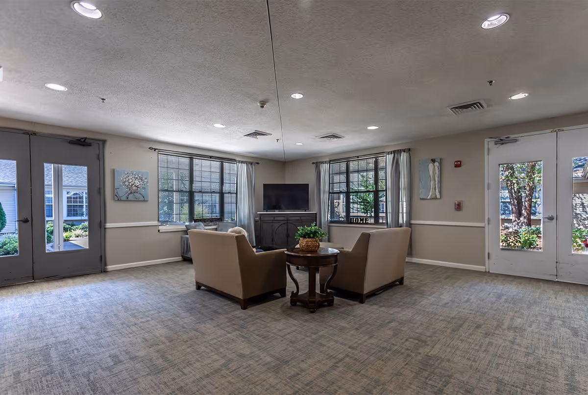 A spacious common area with two beige armchairs facing a round wooden table with a plant on it. Behind the chairs is a TV on a dark wooden stand. The room has large windows with light curtains, beige walls with white trim, and double doors on both sides leading outside. The carpet is light-colored with a subtle pattern, and the ceiling has recessed lighting.
