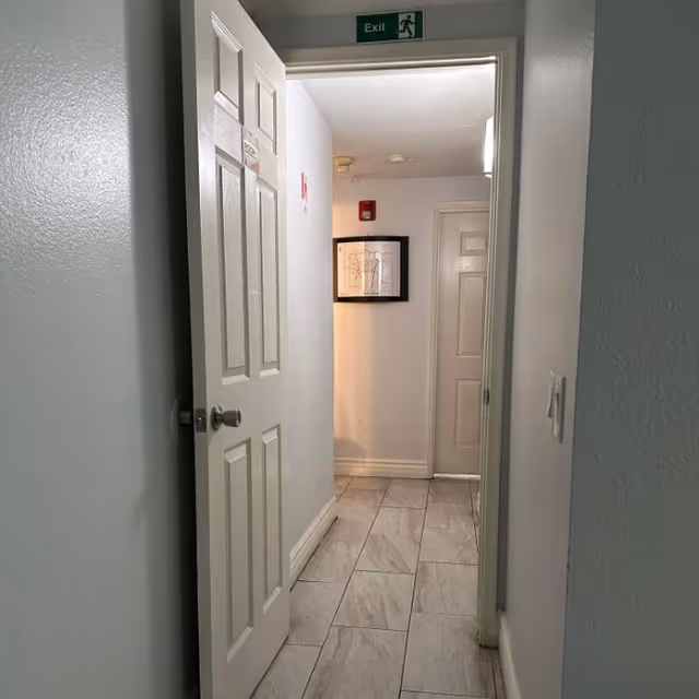 A narrow hallway with light-colored tiled floor and light gray walls. A white door is partially open on the left side, and another closed white door is visible at the end of the hallway. Above the open door is a green exit sign. A framed picture is hanging on the wall at the end of the hallway.